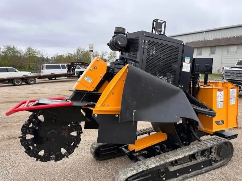 A tracked stump grinder with a black cutting wheel and orange guards parked on a gravel lot